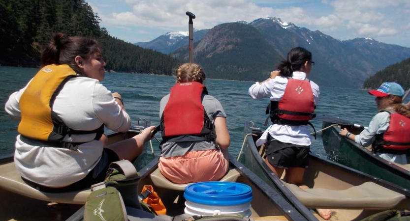 four outward bound students sit at the front of canoes. there are mountains in the background. 
